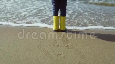 小女孩穿着亮黄色胶靴的特写镜头。孩子站在沙滩上，脚印在沙滩上。